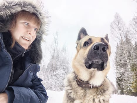 A middle-aged woman takes a cheerful selfie with her German shepherd dog during a winter camping excursion in the snowy landscape.