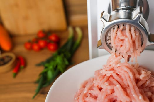 On the left, in a white plate, there is raw pink ground turkey, which comes out of a metal meat grinder on a wooden background next to vegetables and herbs in out-of-focus. The concept of dietary nutrition, healthy and wholesome food, the cooking process. High quality photo