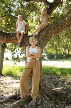 Mother standing by a tree with her arms crossed, smiling while her children sit on a branch above her
