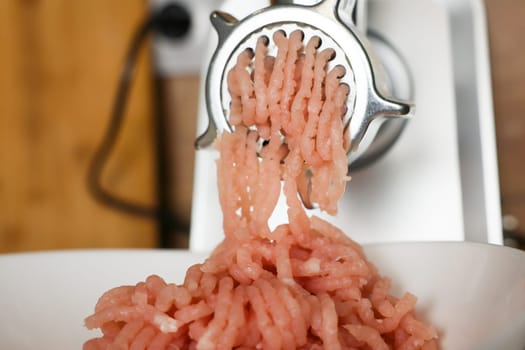 Close-up view: pink minced turkey comes out of a metal meat grinder and sinks into a white plate on a wooden background. The concept of dietary recipes, proper and healthy nutrition, the cooking process. High quality photo