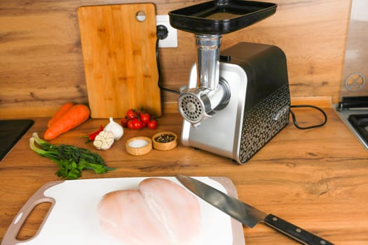 Chicken fillet lies on a white cutting board next to a knife against the background of a wooden kitchen, behind it is a meat grinder, vegetables and spices. The concept of dietary recipes, proper and healthy nutrition, the cooking process. High quality photo