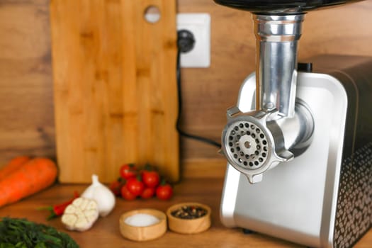 On the right there is an electric metal meat grinder on a wooden background, next to it there are vegetables, spices in wooden bowls and herbs. The concept of cooking, chopping meat. High quality photo