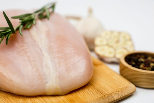 Chicken breast fillet with a sprig of rosemary lies on a wooden board, next to it is a wooden bowl with pepper seasoning and garlic on a white background. The concept of dietary dishes, a recipe for healthy and healthy food. High quality photo