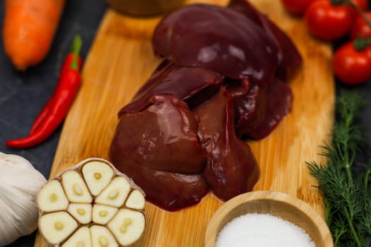 Close-up of raw chicken liver on a wooden plank surrounded by vegetables, seasonings in wooden bowls and herbs on a dark background. The concept of healthy eating, cooking, pate recipe. High quality photo