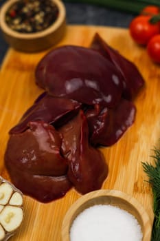 Close-up of raw chicken liver on a wooden board surrounded in out-of-focus garlic, tomatoes, seasonings and salt in wooden bowls and herbs on a dark background. The concept of healthy eating, cooking, pate recipe. High quality photo
