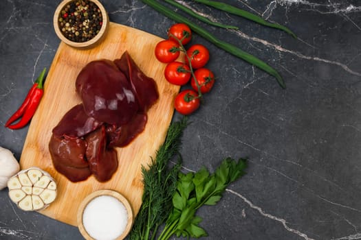 Top view with text space on raw chicken liver on wooden board surrounded by red hot peppers, garlic, tomatoes, seasonings and salt in wooden bowls and herbs on a dark background. The concept of healthy eating, cooking. High quality photo