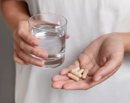 Woman in white t-shirt holding beige capsules in cupped hand and water glass in hand closeup. Female or male taking dietary supplements or vitamins. Healthcare concept