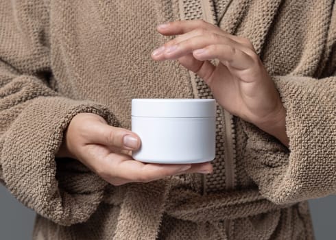 Close up of woman in light brown bathrobe holding white blank cream jar in hands with natural manicure, cosmetic mockup. Bodycare concept. Cropped Image