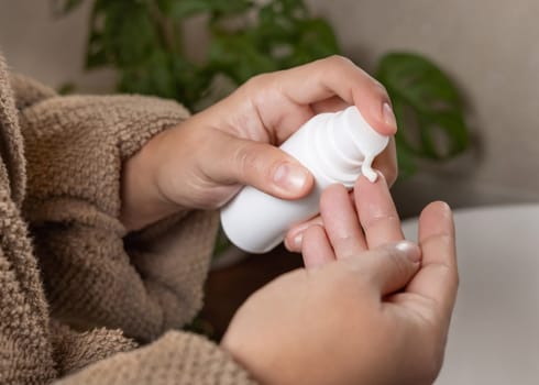 Close up of woman in brown bathrobe pumping cream on finger and holding a cosmetic bottle in other hand in bathroom against green plant and beige wall, beauty routine