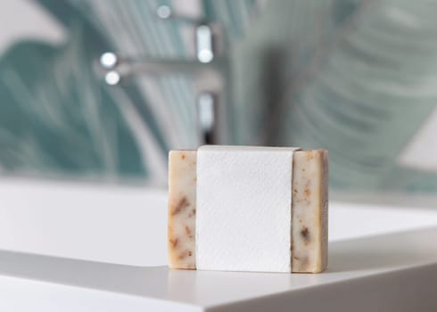 Beige soap bar with blank label on basin near faucet in green tropical bathroom close up, packaging mockup with copy space. Natural plant unbranded product for daily hygiene