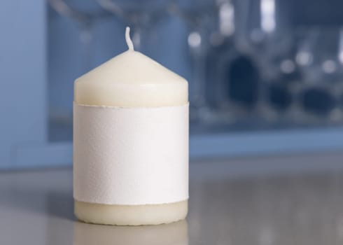 Cream pillar candle with a label on grey kitchen table over blue holder with wine glasses closeup, mockup. Template for advertising and design with copy space