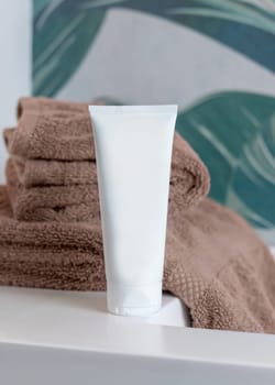 White cream squeeze tube near brown folded towels on basin in green tropical bathroom close up, packaging mockup. Natural plant unbranded cosmetics with copy space. Spa and wellness concept