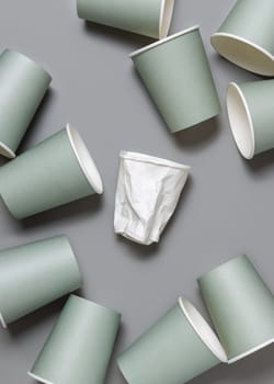 Disposable new light green paper cups and crumpled used white one on gray top view, mockup with copy space. Empty cardboard coffee cups flat lay. Consumerism concept