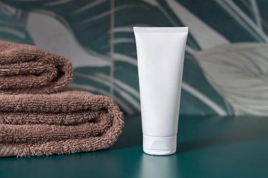White cream squeeze tube near brown folded towels in dark green tropical bathroom close up, packaging mockup. Natural plant unbranded cosmetic container, copy space. Spa and wellness concept