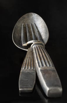 Ancient worn vintage cutlery on the black background. Crossed black metal spoon and fork in dark tone, Highlighting its texture and design, use it as your Space for text, Design, Wallpaper and Poster, Selective focus.