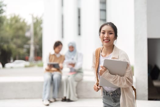 A diverse female student smiling and holding a laptop, with her classmates engaged in a study session in the background at a university campus.