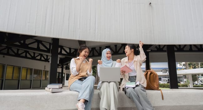 Excited diverse female students celebrating their academic success, raising their hands joyfully while using a laptop and surrounded by books in a university environment.