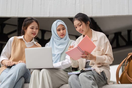 Three diverse female students studying collaboratively outdoors, using a laptop and sharing notes while engaged in their academic work.