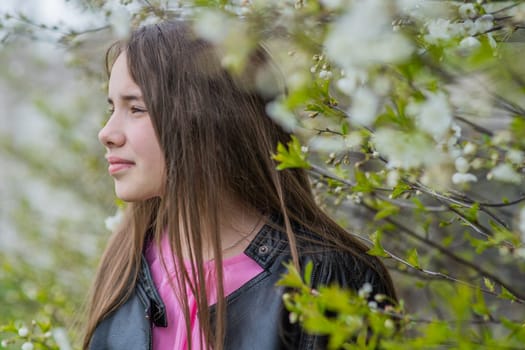 Teenager girl with long brown hair and black leather jacket standing next to blooming tree branches in springtime