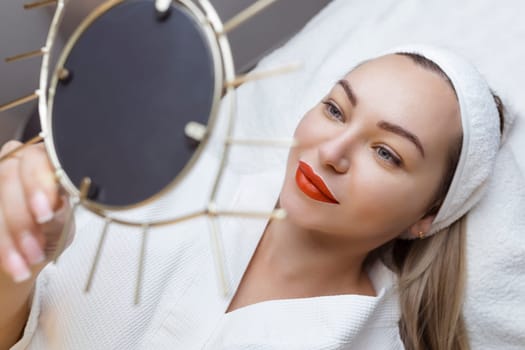 Permanent lip tattooing. A sexy girl with beautiful lips looks into the mirrors lying on the couch at the cosmetologist.