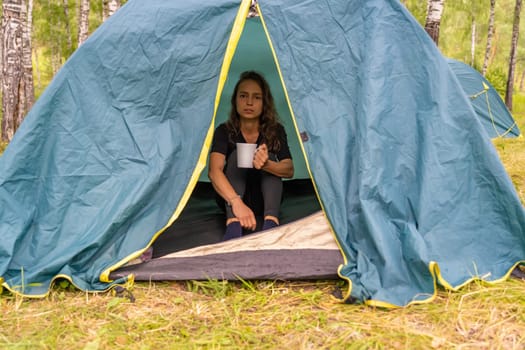 Woman relaxing inside camping tent, drinking warm tea near mountain landscape, experiencing serene outdoor moment with wilderness backdrop