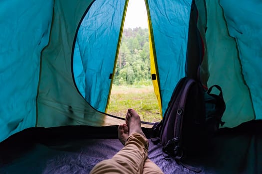 Legs of a camper resting inside a tent, surrounded by a backpack and enjoying a stunning view of a serene forest landscape
