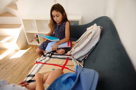 A young girl sits on a sofa, reading books with school supplies beside her in a cozy home environment.