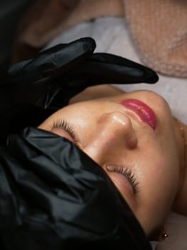 The beautician performs the procedure of microcurrents in black electric gloves to restore facial skin cells. Close-up, vertical