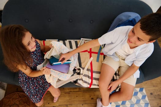 A girl and boy smile while prepping for school, arranging books on a couch. Their playful interaction highlights teamwork.