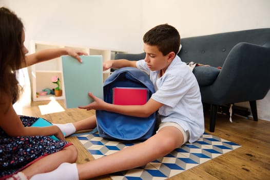 Two children prepare for school by packing notebooks into a school bag, sitting indoors.