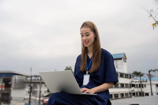 A cheerful female healthcare professional in scrubs smiles while using a laptop outdoors, emphasizing the importance of relaxation and modern technology in her role.
