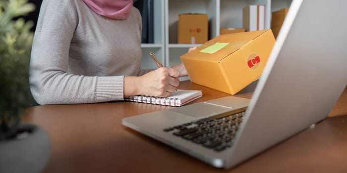 A dedicated Muslim woman engaged in online selling, jotting down notes at her desk, emphasizing the spirit of entrepreneurship in e-commerce.