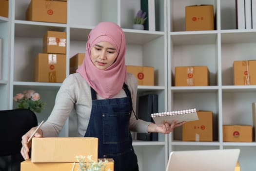 A determined Muslim woman organizing packages and taking notes at her workspace, demonstrating her dedication to managing her online sales effectively.