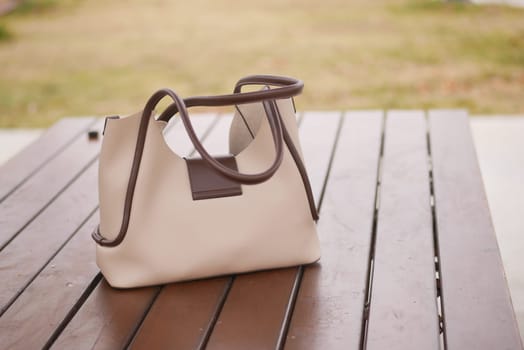 A beige handbag with brown handles rests on a wooden table outdoors.