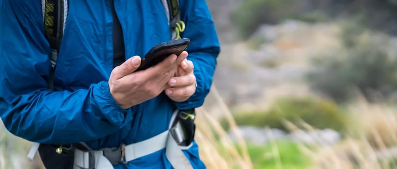 Young man in a turquoise hiking jacket holds a mobile phone in his hands, the beautiful mountains are on the background, a traveler with a backpack walks and uses downloaded maps on his smartphone.