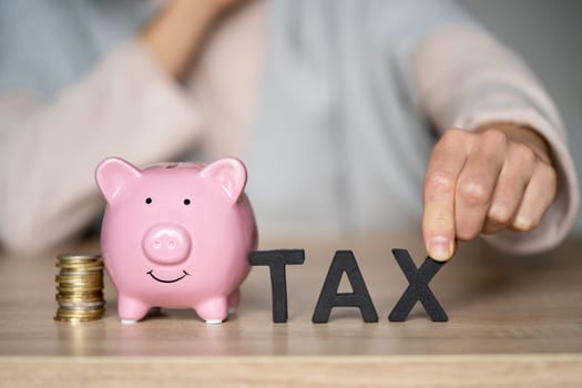 The hand of a young girl puts letters on the table with the words "tax" next to a piggy bank on the table at her workplace, close up view.