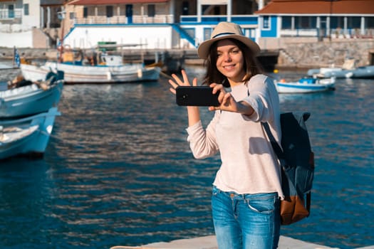 A girl in a hat smiles and takes a selfie in front of a stunning landscape with the sea, yachts and mountains on a sunny day. A woman travels to beautiful places and takes pictures on her smartphone.