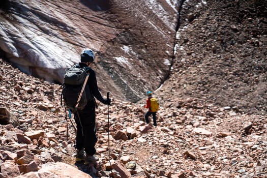 Two people walk in the mountains, hike to the glacier with climbing equipment, in helmets, and with a trekking poles, friends go in for extreme sports and leisure activities together, mountaineering.