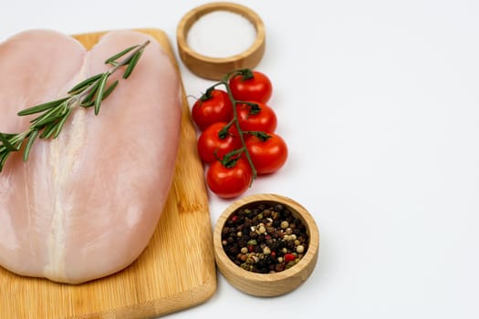 A close-up on the left of a chicken fillet with a sprig of rosemary lies on a wooden cutting board, next to spices and salt in wooden bowls and a sprig of tomatoes on a white background with space for text. The concept of cooking, dietary menu, healthy and natural products. High quality photo