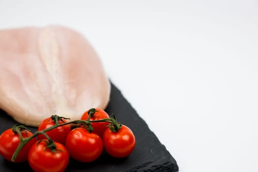 In close-up on the left, chicken fillet with a sprig of tomatoes lies on a black stone board on a white background with space for text. The concept of cooking, dietary menu, healthy and natural products. High quality photo