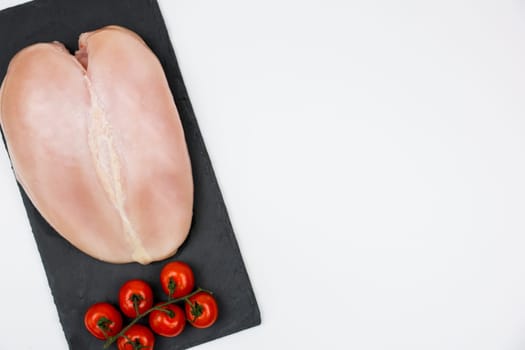 Close-up view from top left: chicken fillet with a sprig of tomatoes next to it lies on a black stone board on a white background with space for text. The concept of cooking, dietary menu, healthy and natural products. High quality photo