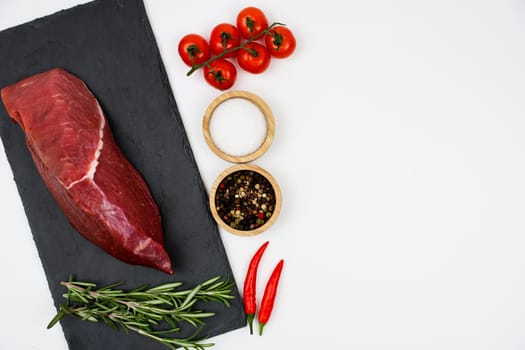 Top view of beef tenderloin on a black stone board with a sprig of rosemary, next to a sprig of tomatoes, spices and salt in wooden bowls and red hot peppers on a white background with space for text. The concept of cooking, dietary menu, healthy and natural products. High quality photo