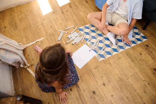 Two children enjoying creative activities, drawing with markers on a wooden floor. The scene captures teamwork and artistic expression.