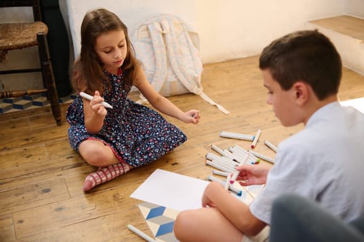 Two children are seated on the wooden floor, engaging in creative drawing activities using markers, showcasing their colors and imagination.