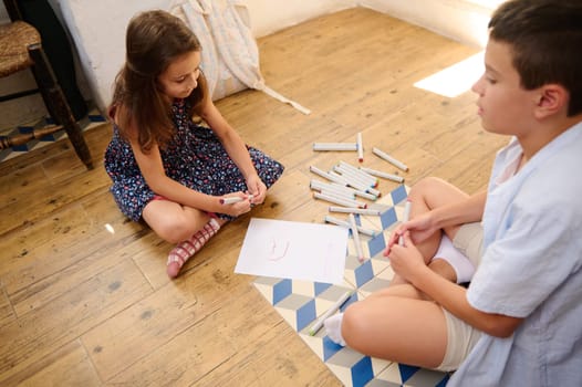 Two children sit on the floor drawing on paper with markers. They engage in creative activities, surrounded by art supplies, showcasing childhood imagination and companionship.