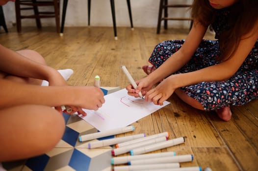 Children drawing together on paper using markers, engaging in creativity and fun on a wooden floor.