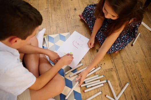 Two children sitting on a wooden floor, drawing on paper with colorful markers, surrounded by art supplies, fostering creativity and collaboration in a fun and artistic setting.