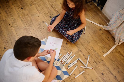 Two children sitting on a wooden floor, using markers and paper for sketching. The image captures creativity, childhood bonding, and indoor leisure. Art supplies scattered around enhance the creative atmosphere.