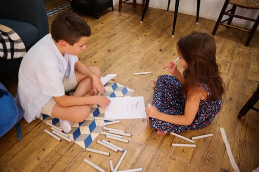 Two children are sitting indoors drawing with colorful markers on paper, fostering creativity and collaboration.