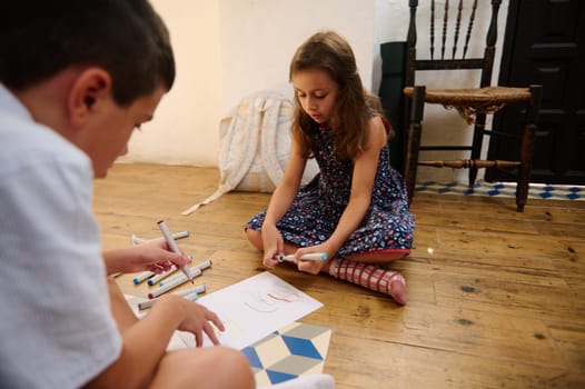 Two children are enjoying creative activities, drawing on paper with markers, sitting on a wooden floor in a cozy indoor environment. This captures childhood innocence and artistic expression.